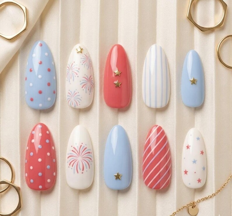 May include: A collection of ten artificial nails in various designs. The nails feature red, white, and blue colors with patterns like polka dots, stripes, fireworks, and star accents. The nails are oval-shaped and arranged on a light-colored surface.
