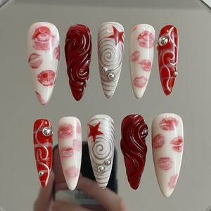以下が含まれることがあります： 10本のアーモンド型の長い人工爪のセット。デザインには、白地に赤いキスのプリント、渦巻き模様、赤い星、銀色の装飾が含まれます。爪は反射面に配置されています。
