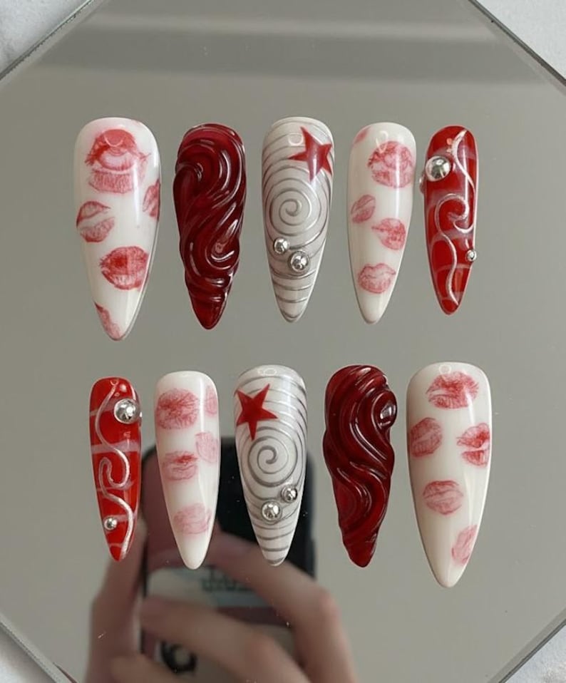 以下が含まれることがあります： 10本のアーモンド型の長い人工爪のセット。デザインには、白地に赤いキスのプリント、渦巻き模様、赤い星、銀色の装飾が含まれます。爪は反射面に配置されています。
