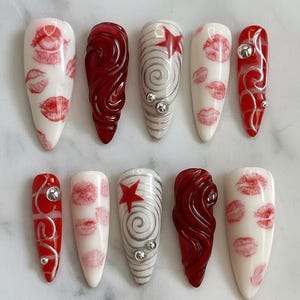 以下が含まれることがあります： さまざまなデザインの人工爪10本セット。赤い唇のプリントが施された白い爪、シルバーの渦巻きと宝石が付いた赤い爪、赤い星とシルバーの渦巻き模様が特徴の爪があります。