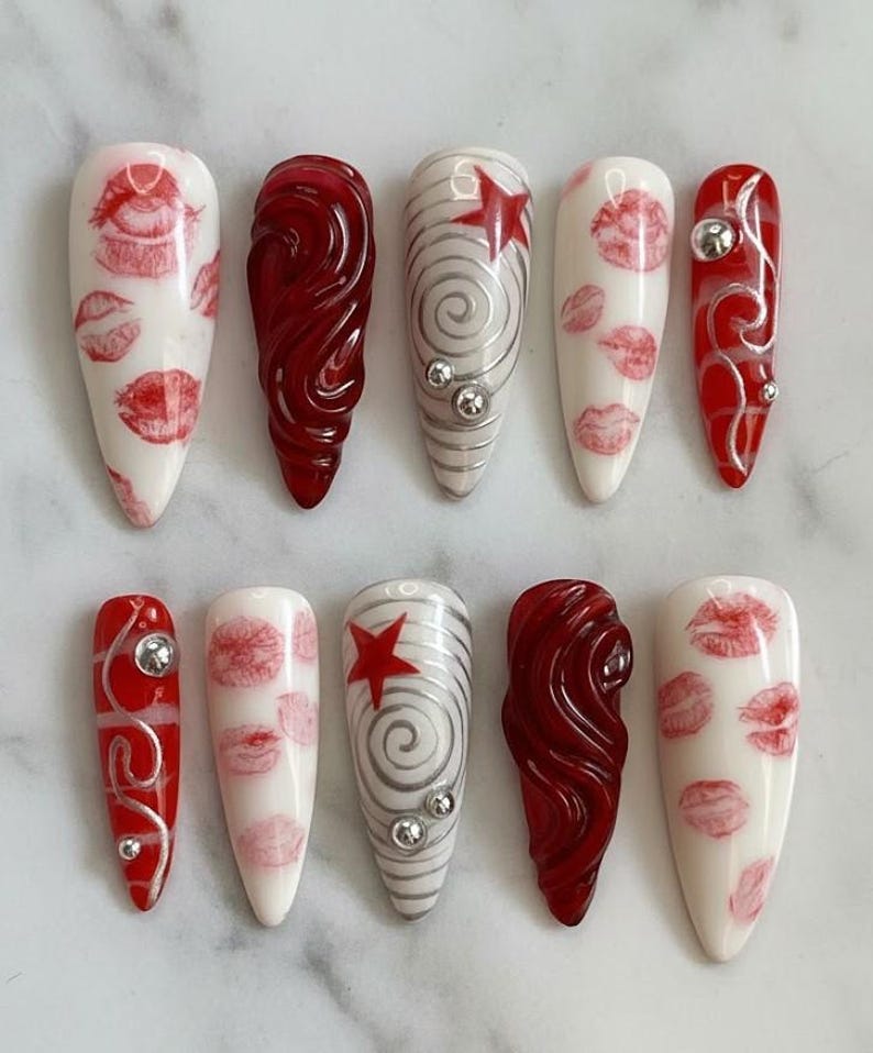 以下が含まれることがあります： さまざまなデザインの人工爪10本セット。赤い唇のプリントが施された白い爪、シルバーの渦巻きと宝石が付いた赤い爪、赤い星とシルバーの渦巻き模様が特徴の爪があります。
