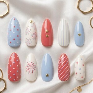 May include: A set of ten almond-shaped false nails with various designs. The designs include red and white polka dots, fireworks, stripes, and stars. Some nails feature gold star embellishments. The color palette includes red, white, and blue.
