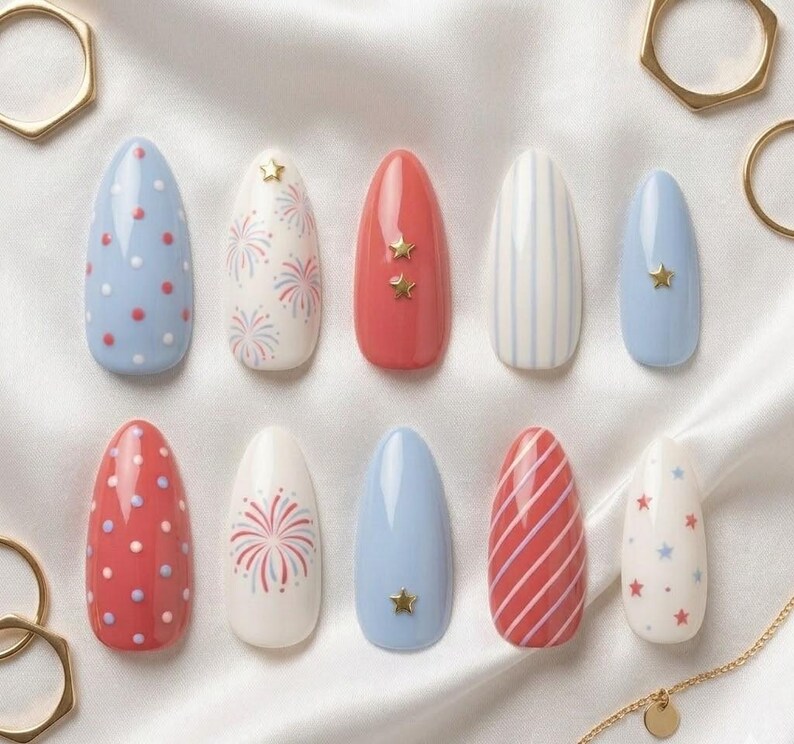 May include: A set of ten almond-shaped false nails with various designs. The designs include red and white polka dots, fireworks, stripes, and stars. Some nails feature gold star embellishments. The color palette includes red, white, and blue.