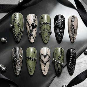 May include: A set of ten almond-shaped press-on nails featuring olive green, black, and cream colors. The designs include raised black stitching, silver metal bolt accents, a black heart, a spider web, and marble patterns.