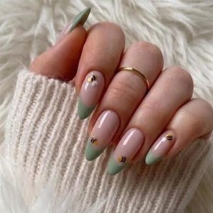 May include: A hand with long, almond-shaped nails. The nails feature a light pink base with a green French tip. Each nail has a small bee design with gold accents. A gold ring is on one finger.