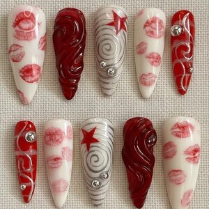 以下が含まれることがあります： 10本のアーモンド型の長い人工爪のセット。デザインには、白地に赤い唇、渦巻く赤いデザイン、赤い星が付いたシルバーの渦巻き、シルバーのアクセントが含まれます。爪はニュートラルな背景に配置されています。