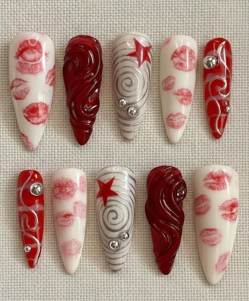 以下が含まれることがあります： 10本のアーモンド型の長い人工爪のセット。デザインには、白地に赤い唇、渦巻く赤いデザイン、赤い星が付いたシルバーの渦巻き、シルバーのアクセントが含まれます。爪はニュートラルな背景に配置されています。