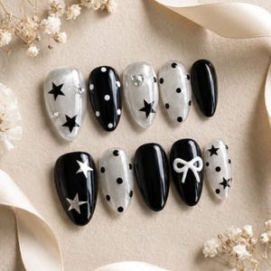 May include: A set of ten almond-shaped press-on nails featuring a black and translucent white color palette. The designs include black stars, white polka dots, silver star accents, and a raised white bow detail on a glossy black base finish.