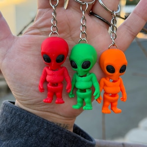 May include: Three alien-shaped keychains in red, green, and orange. Each keychain features a small, humanoid alien figure with large black eyes and a silver chain. The figures are made of a smooth, solid material.