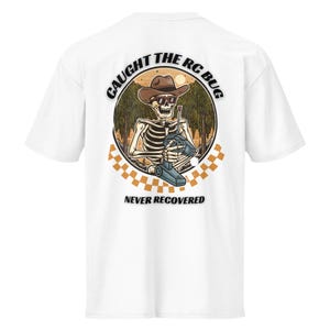 Peut inclure: T-shirt blanc avec un motif d'un squelette portant un chapeau de cowboy et des lunettes de soleil, tenant une manette RC. Le motif est dans un cadre circulaire avec le texte "CAUGHT THE RC BUG" et "NEVER RECOVERED". Un motif à carreaux est en bas.