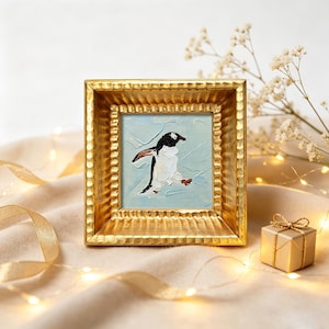 Può includere: Un piccolo dipinto a olio quadrato raffigurante un pinguino bianco e nero su sfondo azzurro, racchiuso in una cornice dorata testurizzata con un bordo smussato profondo. L'opera mostra pennellate visibili e una spessa consistenza pittorica.