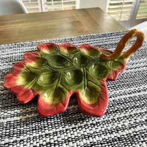 Puede incluir: Un plato de servicio de cerámica con forma de hoja de roble grande y un asa curva. La superficie presenta un acabado brillante con un degradado de verde bosque intenso en el centro que pasa a rosa rojizo en los bordes, con nervaduras en relieve.