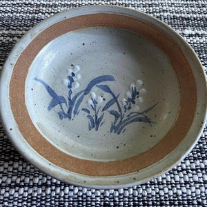 May include: A round ceramic bowl with a light blue interior and a brown rim. The bowl features a hand-painted floral design in blue and white. The bowl is set against a black and white patterned background.