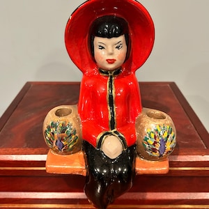 Puede incluir: Una figura de cerámica de una persona con chaqueta roja con ribete negro y un gran sombrero circular rojo. La figura está sentada en un banco, sosteniendo dos recipientes esféricos de color tostado, decorados con un motivo de plumas de pavo real.