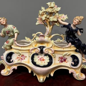 Könnte beinhalten: Antikes Porzellan-Mittelstück in Creme, Rosa und Marineblau. Es zeigt zwei Figuren, einen Baum und florale Designs. Die Figuren tragen detaillierte Kleidung. Dieses dekorative Objekt ist wahrscheinlich für die Präsentation gedacht.