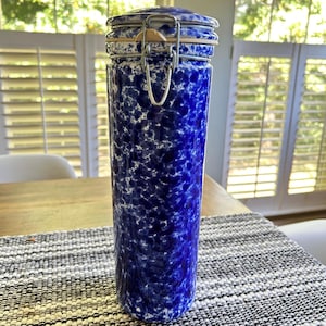 May include: A tall, cylindrical canister featuring a blue and white speckled pattern. The container includes a matching lid secured by a metal wire bail closure mechanism with a rubber gasket seal visible at the top rim of the vessel.