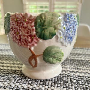 以下が含まれることがあります： ピンクとブルーの紫陽花の花と緑の葉が立体的なレリーフとして施された白い陶器製のボウル。ボウルの表面には繊細なひび割れ模様の釉薬がかけられており、小さな円形の台座の上に置かれています。