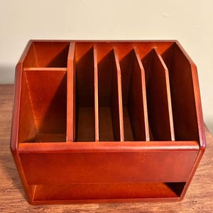 May include: A medium-brown wooden desk organizer featuring a solid front panel, a single open compartment on the left side, and four vertical dividers creating five narrow slots for storing documents, mail, or stationery items on the right side.