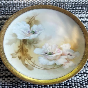 May include: A round, antique porcelain plate with a gold rim and a floral design. The plate features hand-painted white flowers with pink and green accents on a cream-colored background. The plate has a gold border.
