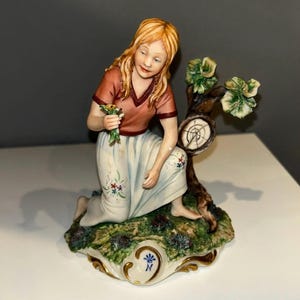 May include: A porcelain figurine of a kneeling figure with long blonde hair, wearing a red top and white skirt with floral accents. The figurine holds flowers and is next to a tree with a clock design.