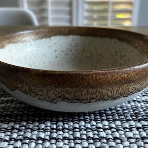 Puede incluir: Un cuenco de cerámica con interior blanco moteado y exterior marrón texturizado. El borde del cuenco es de un marrón más oscuro, creando contraste con el interior más claro. El cuenco está sobre una superficie con estampado en blanco y negro.