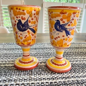 May include: Two ceramic goblets featuring a hand-painted design of a blue bird perched among yellow and orange floral patterns. The stems and circular bases are decorated with horizontal yellow and orange stripes and scalloped red line details.