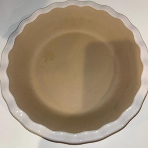 Może przedstawiać: Okrągła forma do ciasta z kremowym wnętrzem i białym, falistym brzegiem. Naczynie jest puste i wydaje się być wykonane z ceramiki lub kamionki. Projekt jest prosty i klasyczny, odpowiedni do pieczenia ciast i innych deserów.