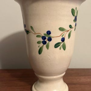 May include: A cream-coloured ceramic vase with a speckled finish and a flared rim. The vase features a hand-painted design of thin brown branches with green leaves and small dark blue berries encircling the upper portion of the vessel body.