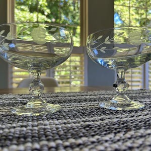 May include: Two clear glass coupe glasses featuring a delicate etched floral and vine pattern around the bowl. Each glass has a short, multi-faceted stem and a circular base, designed for serving beverages with a wide, shallow bowl shape.