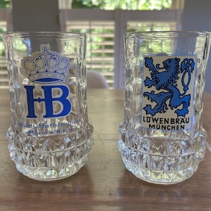 May include: Two clear glass beer mugs with textured dimpled bases. The left mug features a blue crown logo with the text HB Staatliches Hofbräuhaus München. The right mug displays a blue lion emblem with the text Löwenbräu München in a rectangular frame.