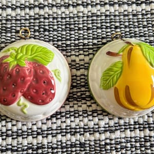 May include: Two round ceramic wall plaques featuring embossed fruit designs. One plaque displays a pair of red strawberries with green leaves, while the other shows a yellow pear with green leaves. Each piece includes a small metal hanging loop.