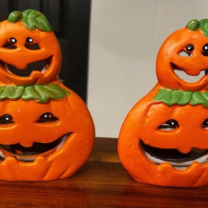 May include: Two orange ceramic pumpkin figurines, each featuring a smaller pumpkin stacked on top of a larger one. Both pumpkins have carved, smiling faces with cut-out eyes and mouths, and green painted leaves at the base of the top pumpkin.