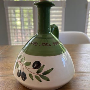 May include: A ceramic olive oil bottle with a green neck and handle, and a white body. The bottle is decorated with a painted olive branch and the words "ORI DEL FRA". The bottle has a wooden stopper.