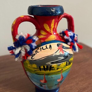May include: A small red ceramic amphora vase with two handles, featuring a hand-painted landscape scene with the text "SICILIA" and a mountain. The vase is decorated with yellow floral accents and two fluffy pom-poms in red, white, and blue yarn.