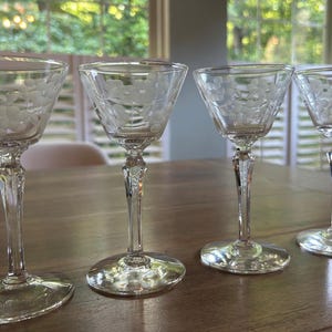 May include: A set of four clear glass stemmed glasses, each featuring a delicate etched pattern of grapes and vine leaves around the bowl. The glasses have a slender stem and a circular base, providing a consistent design across all four pieces.