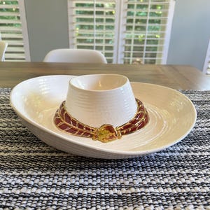 May include: A cream-colored ceramic serving dish shaped like a sombrero hat. The wide brim serves as a bowl, while the crown forms a central cup. A decorative band in deep red with gold-toned trim encircles the base of the crown.