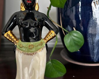 Może przedstawiać: Ceramiczna figurka kobiety o czarnej skórze, noszącej białe nakrycie głowy z klejnotami, złote kolczyki oraz zielono-złoty pas. Figurka ma ręce na biodrach i nosi kremową spódnicę.