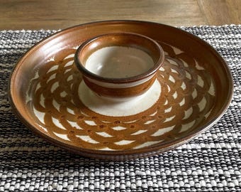 May include: A round ceramic serving dish featuring a central bowl attached to a wider plate. The piece has a cream-colored base with a repeating brown, scale-like pattern radiating outward from the center. The rim and bowl edge are brown.