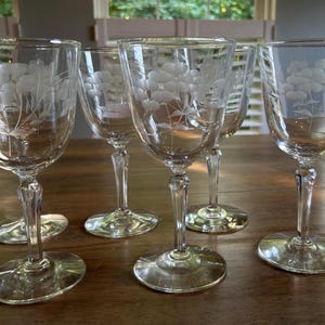 May include: A set of five clear glass wine glasses, each featuring a delicate etched floral pattern on the bowl. The glasses have a slender stem and a circular base, showcasing a transparent design with intricate white floral engravings.