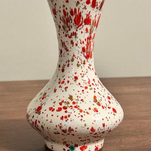 Puede incluir: Un jarrón de cerámica con base blanca que presenta un patrón moteado de salpicaduras de pintura roja brillante y dorada metálica. El jarrón tiene un cuerpo ancho y redondeado que se estrecha hacia un cuello delgado y un borde acampanado.