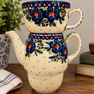 Puede incluir: Un juego de té individual de cerámica compuesto por una tetera y una taza apilada encima. La pieza de color crema presenta un patrón pintado a mano de flores azul oscuro con centros naranjas, hojas verdes y pequeños puntos azules por toda la superficie.