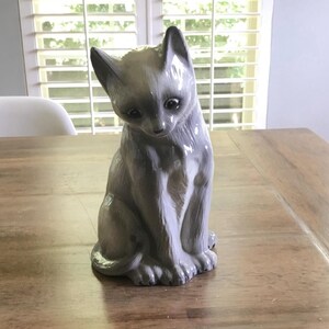 May include: A glossy ceramic figurine of a seated cat with a light gray and white coat. The cat has large dark eyes, pointed ears, and a tilted head posture. The surface of the figurine is smooth and reflective, capturing light across its curved form.