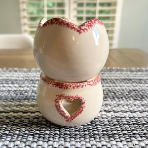 May include: A two-piece ceramic wax warmer featuring a cream-colored base and top. The top bowl has a scalloped edge with red speckled detailing. The base includes a heart-shaped cutout and matching red speckled trim around the top rim.