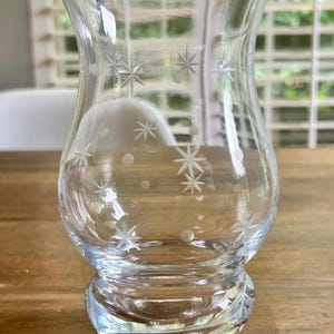 May include: A clear glass hurricane candle holder with a flared rim and a rounded base. The exterior surface features an etched pattern consisting of multiple eight-pointed starburst designs interspersed with small circular dots throughout.