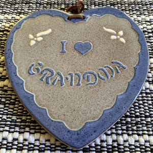Könnte beinhalten: Eine herzförmige Keramikplakette mit gesprenkeltem beigem Zentrum und blauem Rand. Der Text "I [Herzsymbol] GRANDMA" ist in Blau eingeprägt, begleitet von zwei kleinen weißen Blumenakzenten. Ein braunes Lederband ist an der Oberseite befestigt.