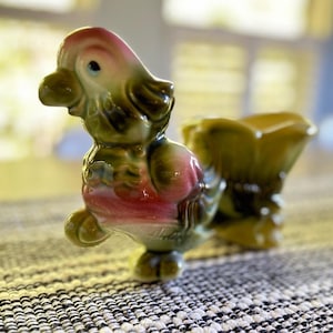 May include: A ceramic bird figurine featuring a glossy finish with blended olive green and soft pink coloration. The bird is depicted in a standing position with a small, open-topped container attached to its back, showcasing a smooth, glazed surface.