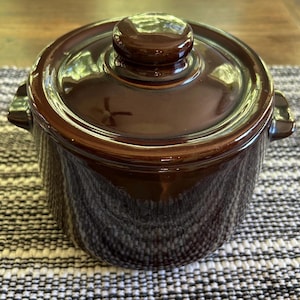 May include: A small, brown ceramic casserole dish with a lid. The dish has a glossy finish and two small handles. The lid has a rounded knob. The dish is on a patterned surface.
