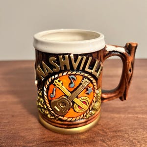 Könnte beinhalten: Ein brauner Keramikbecher mit weißem Rand und einem Henkel in Form eines Baumastes. Auf der Vorderseite steht das Wort NASHVILLE in erhabenen Buchstaben über einem kreisförmigen Emblem mit Gitarre, Banjo und Noten auf orangefarbenem Grund.