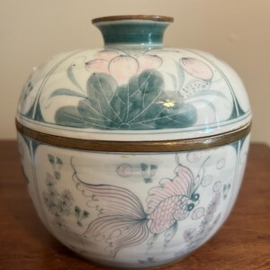 May include: A round ceramic lidded jar featuring a hand-painted design of pink goldfish and green aquatic plants. The container has a brass-colored metal rim where the lid meets the base and a small, circular knob handle on top of the lid.