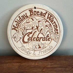 May include: A round, off-white ceramic bread warmer featuring brown embossed text and illustrations. The text reads "Holiday Bread Warmer," "Celebrate," and "Family & Friends." The design includes grape clusters, leaves, and a loaf of bread.